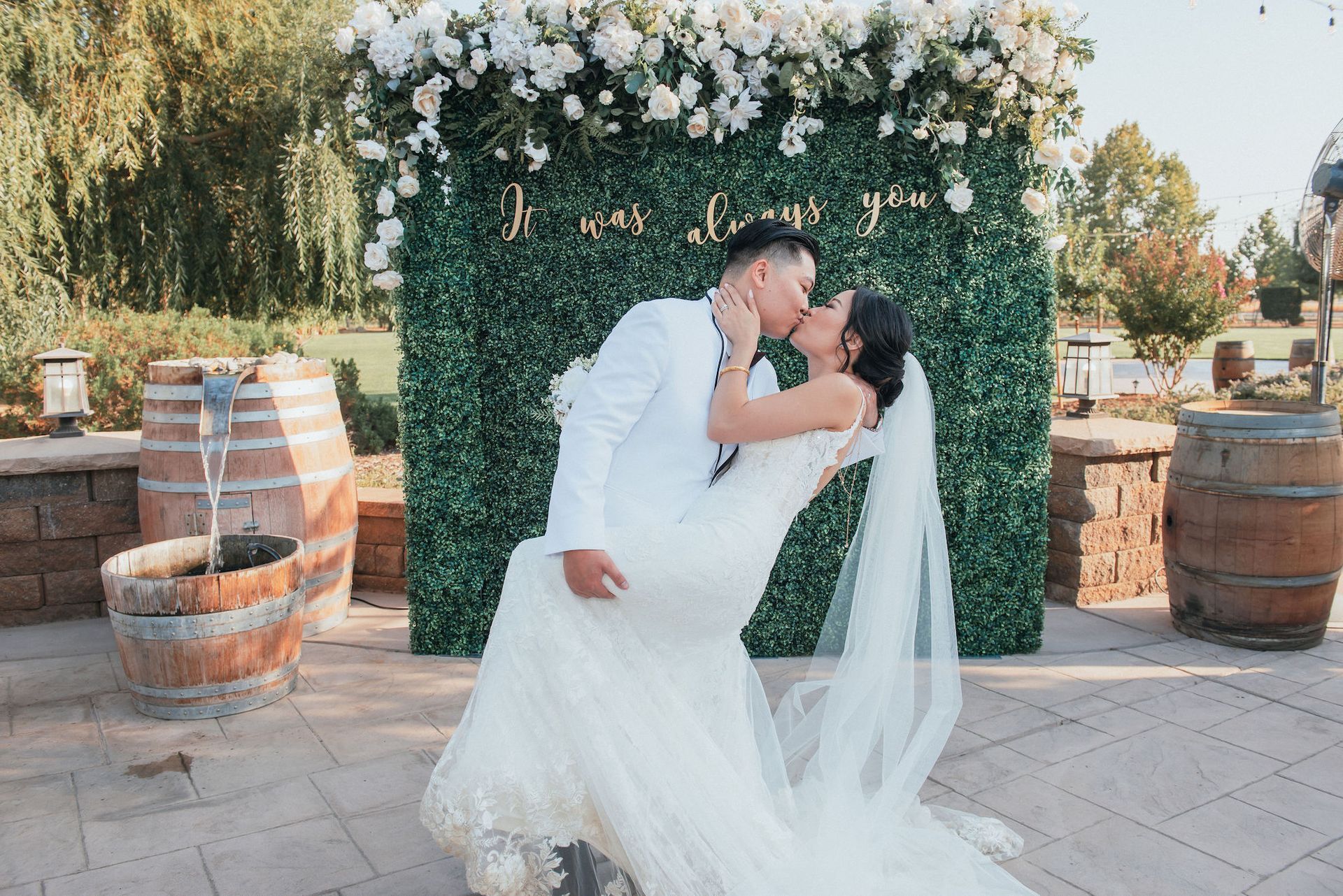 A bride and groom are kissing in front of a wall of flowers.