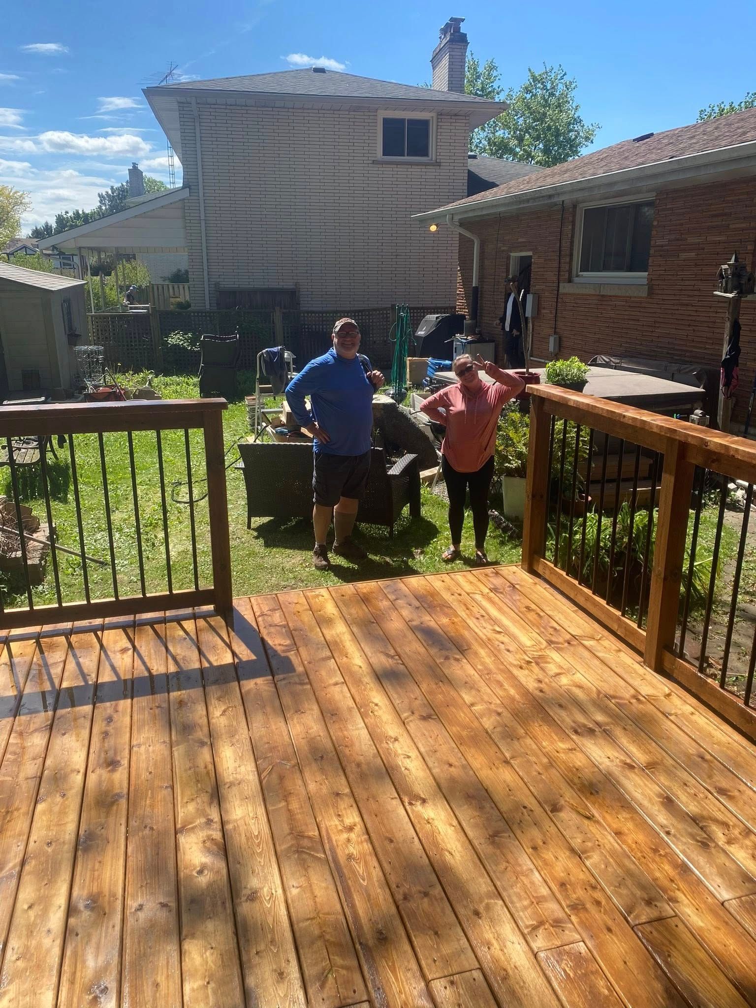 Two people stand in a backyard, near a wooden deck. The sky is blue.