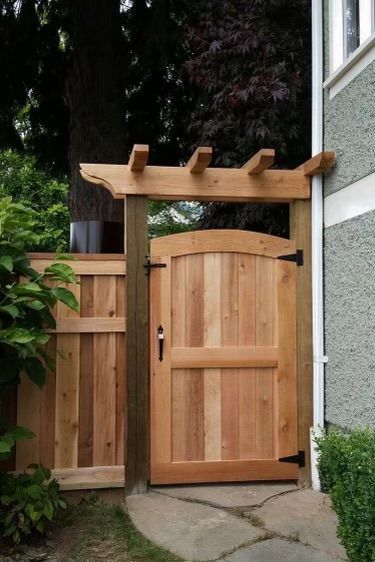 Wooden gate with pergola over a stone path, leading to a house, surrounded by greenery.