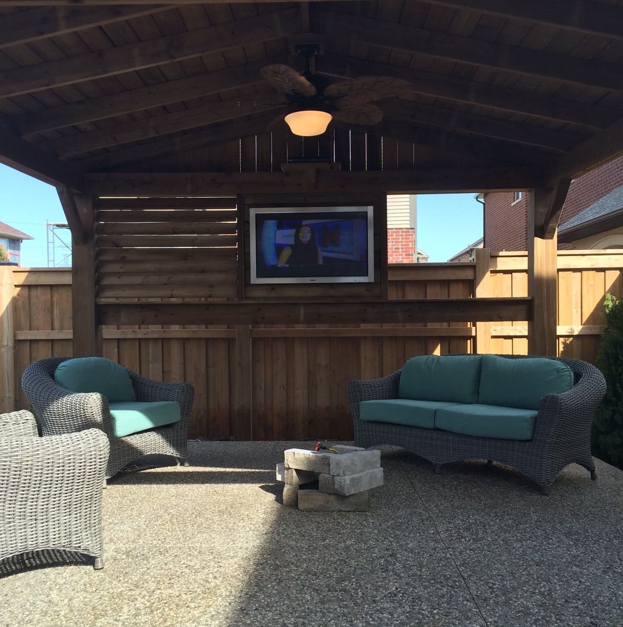 Outdoor patio with wicker furniture, a mounted TV, and a ceiling fan under a wooden structure.