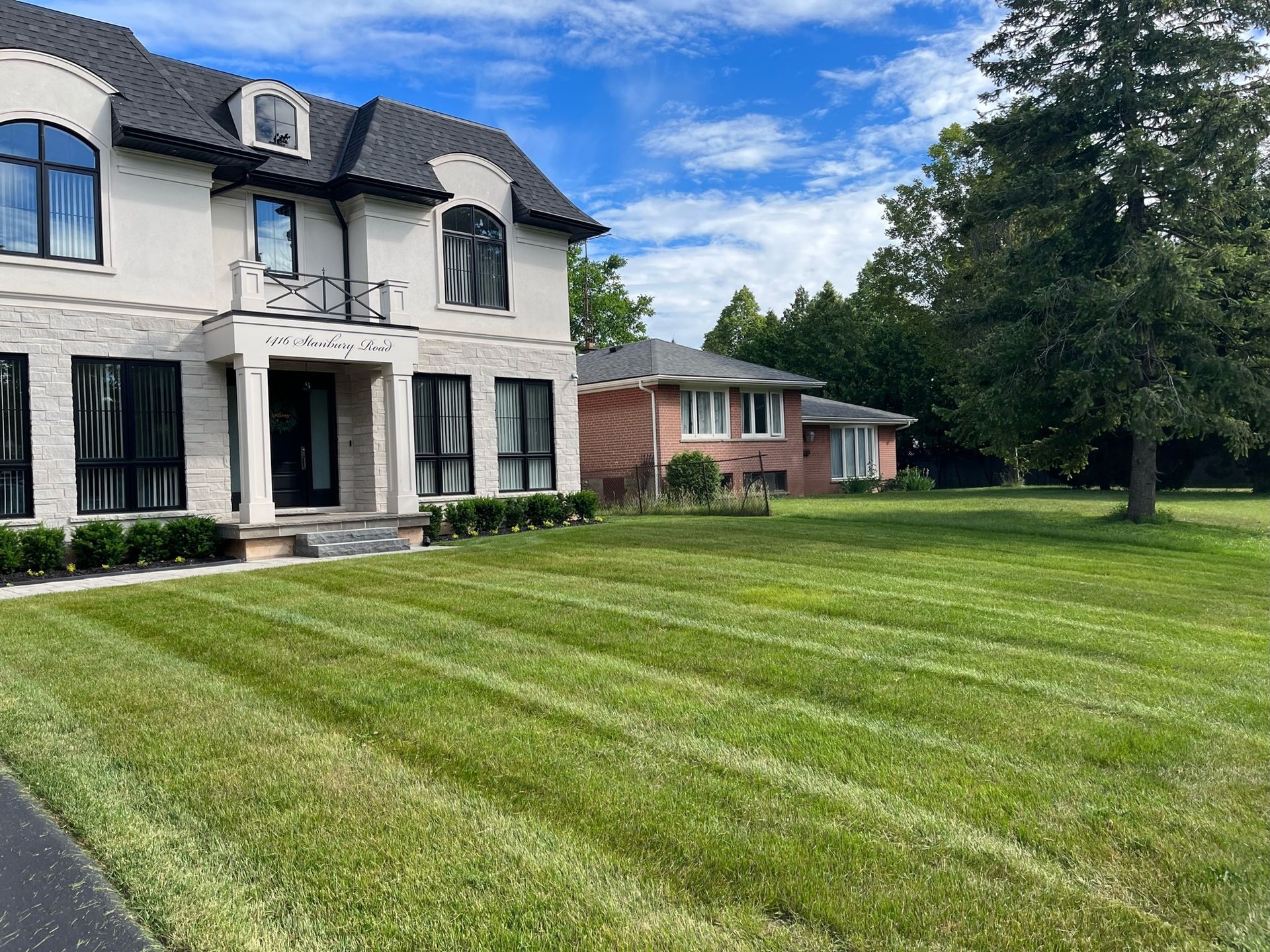 Mowed green lawn in front of two houses, one large with stone and the other brick, under a blue sky.