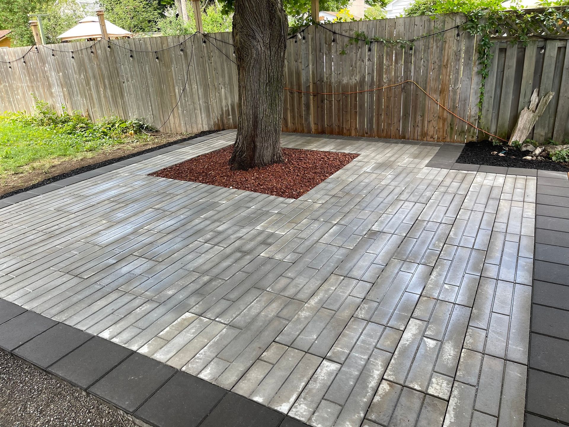 Brick patio around a tree in a backyard with a wooden fence.