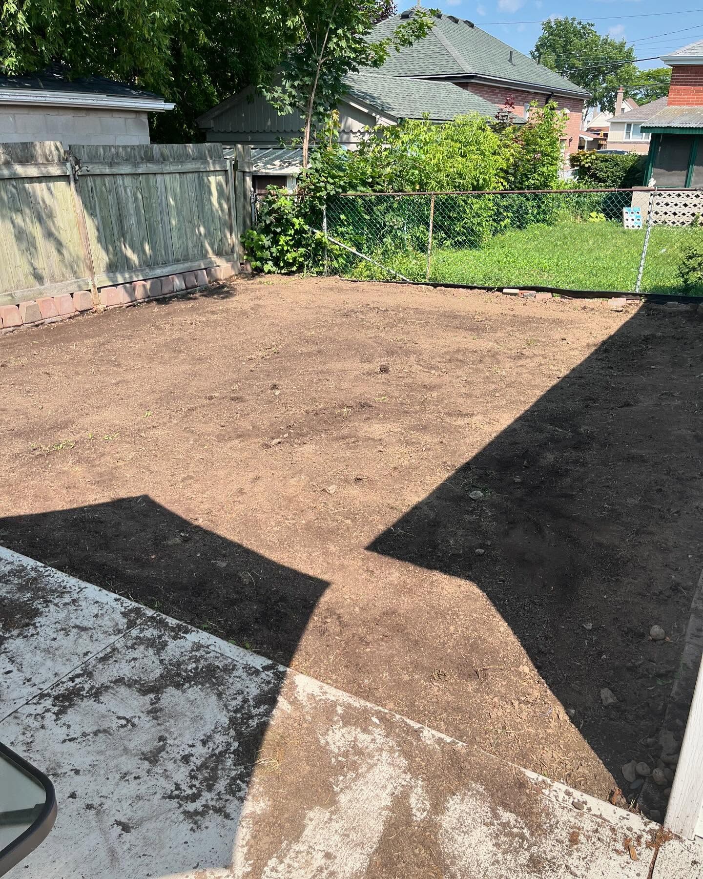 A cleared backyard, mostly dirt. Concrete patio in the foreground, green fence, grass, and houses in the background.