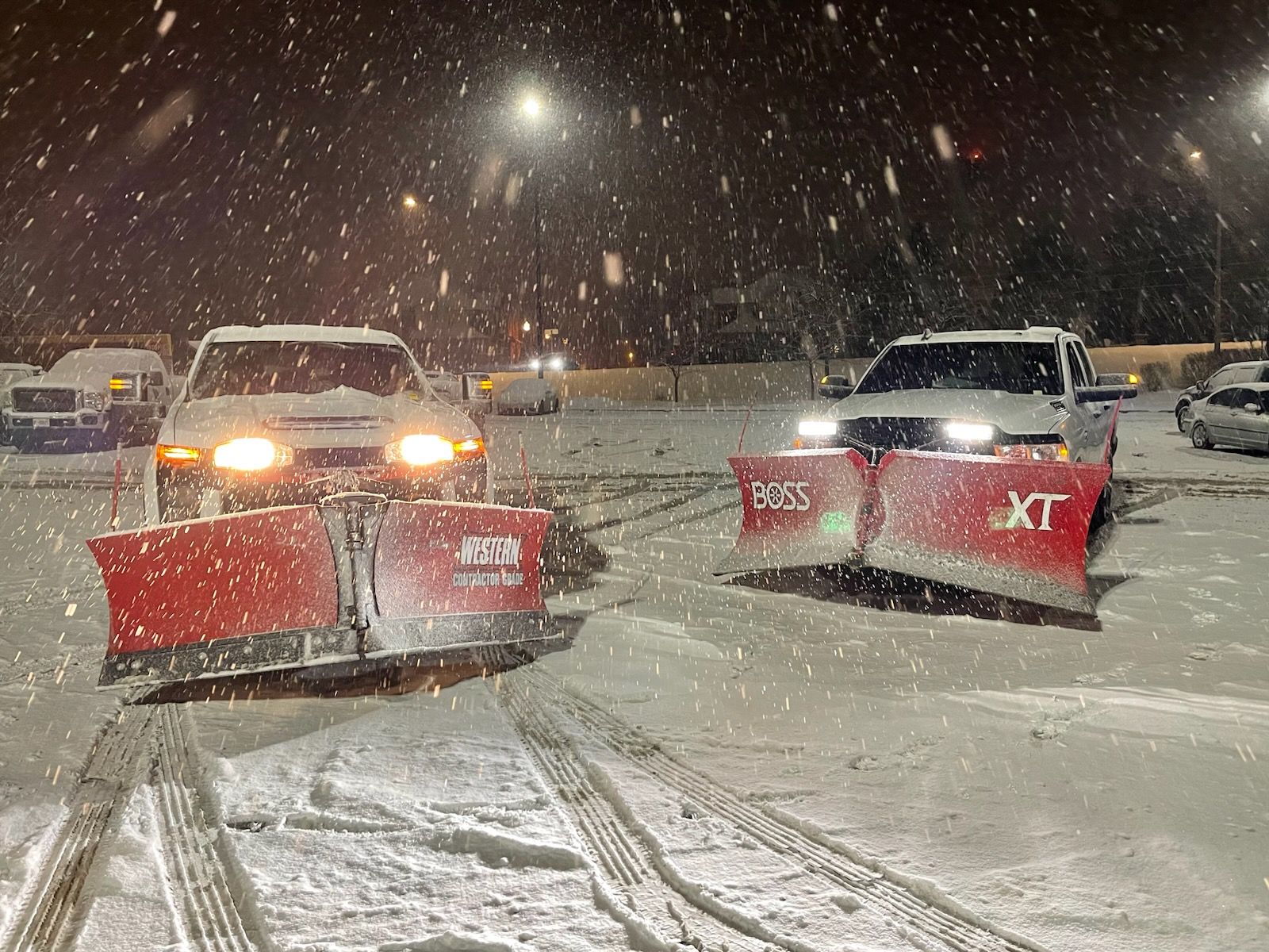 Two snowplows clearing a snowy parking lot at night.