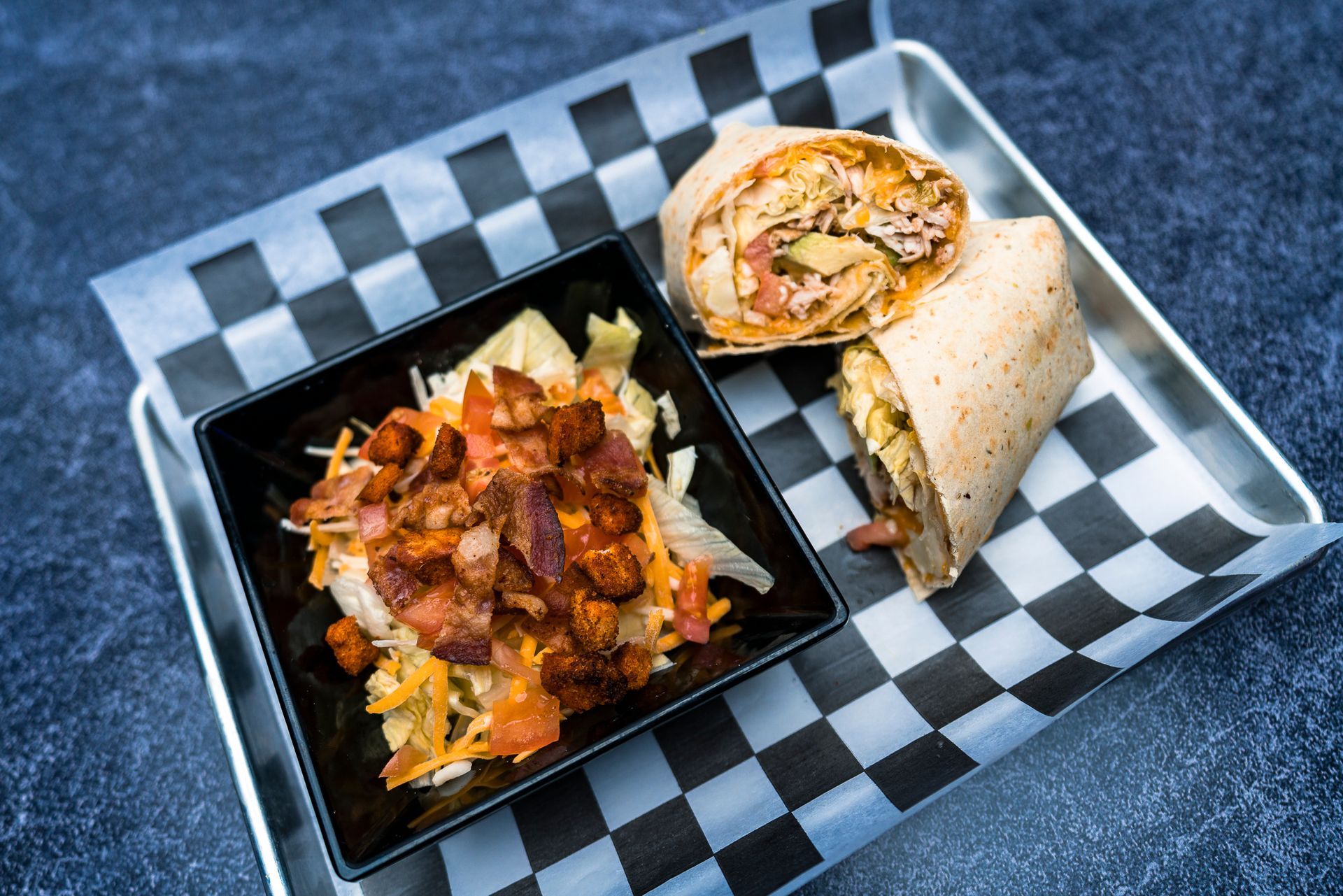 A burrito and a bowl of food on a checkered paper on a tray.