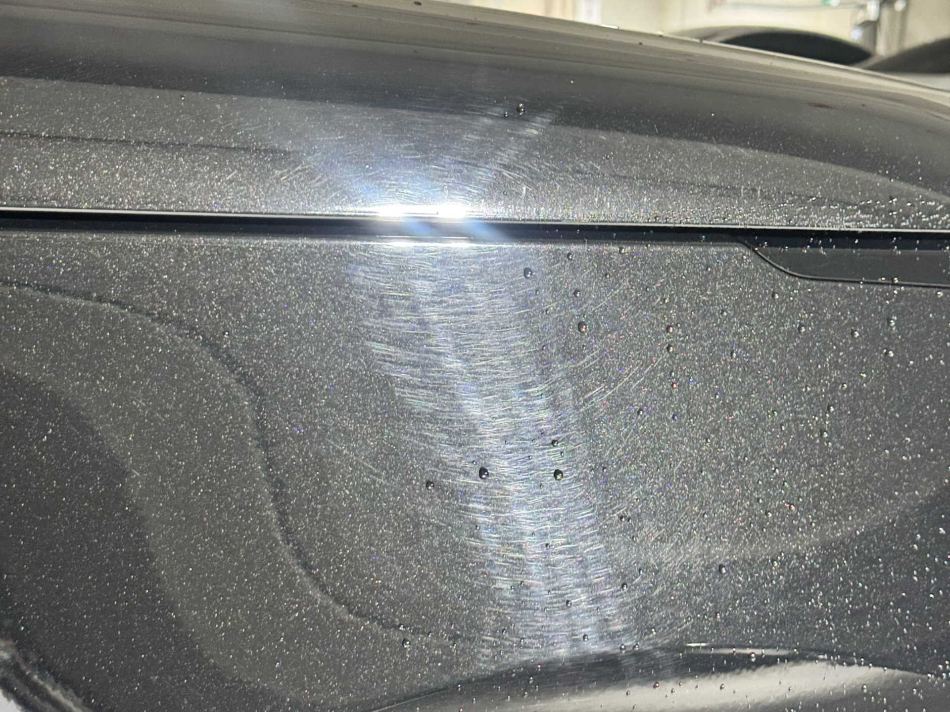Close-up of a dark gray car hood showing swirl marks and water droplets.