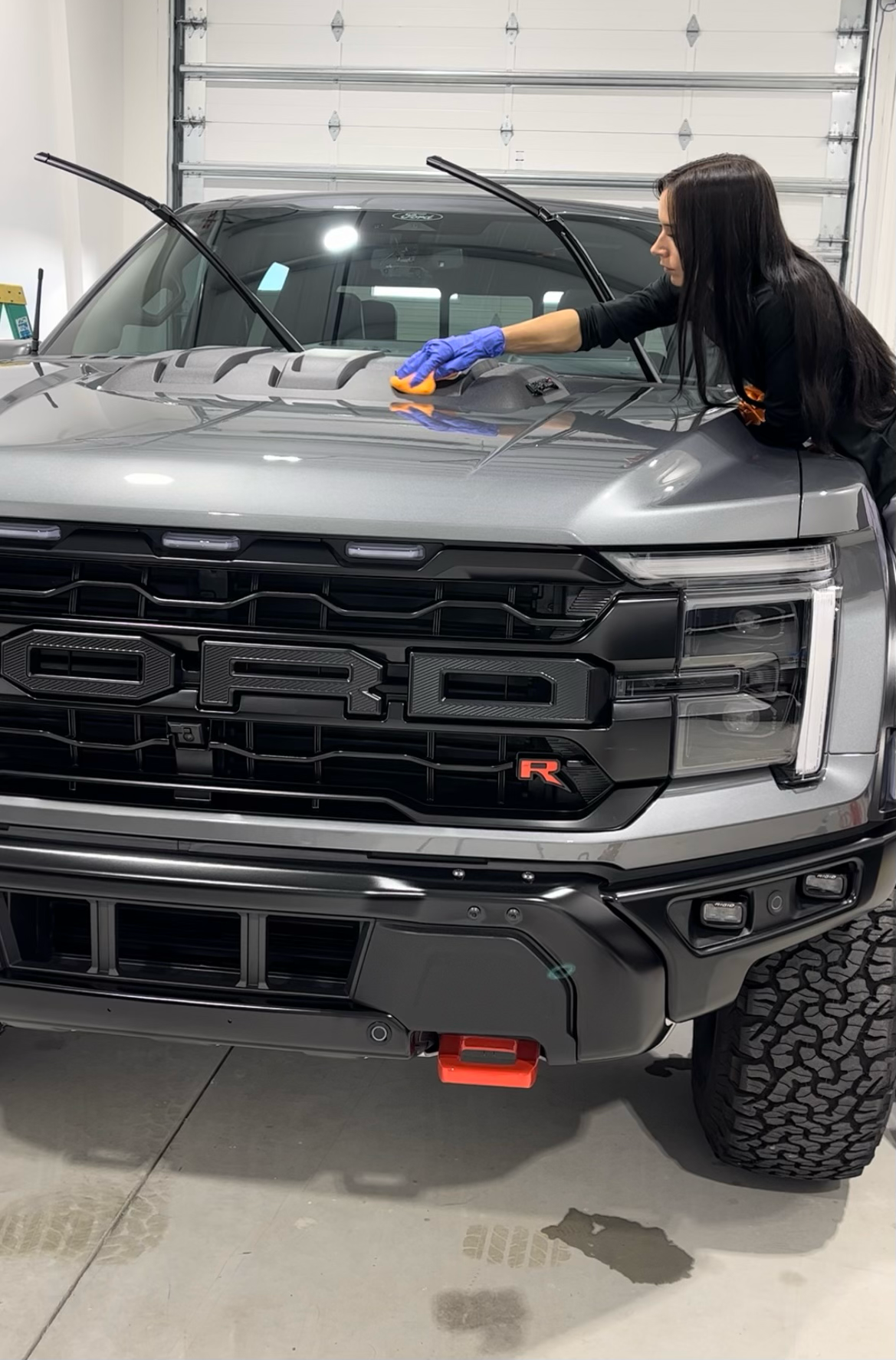 Woman washing a gray Ford Raptor truck hood with a sponge, inside a garage.