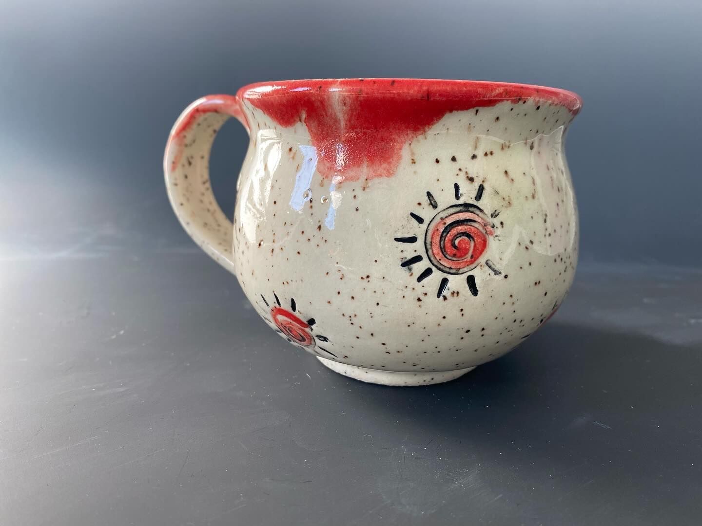 Ceramic mug with a red-glazed rim and sun design. It has a rounded body and a speckled white base.