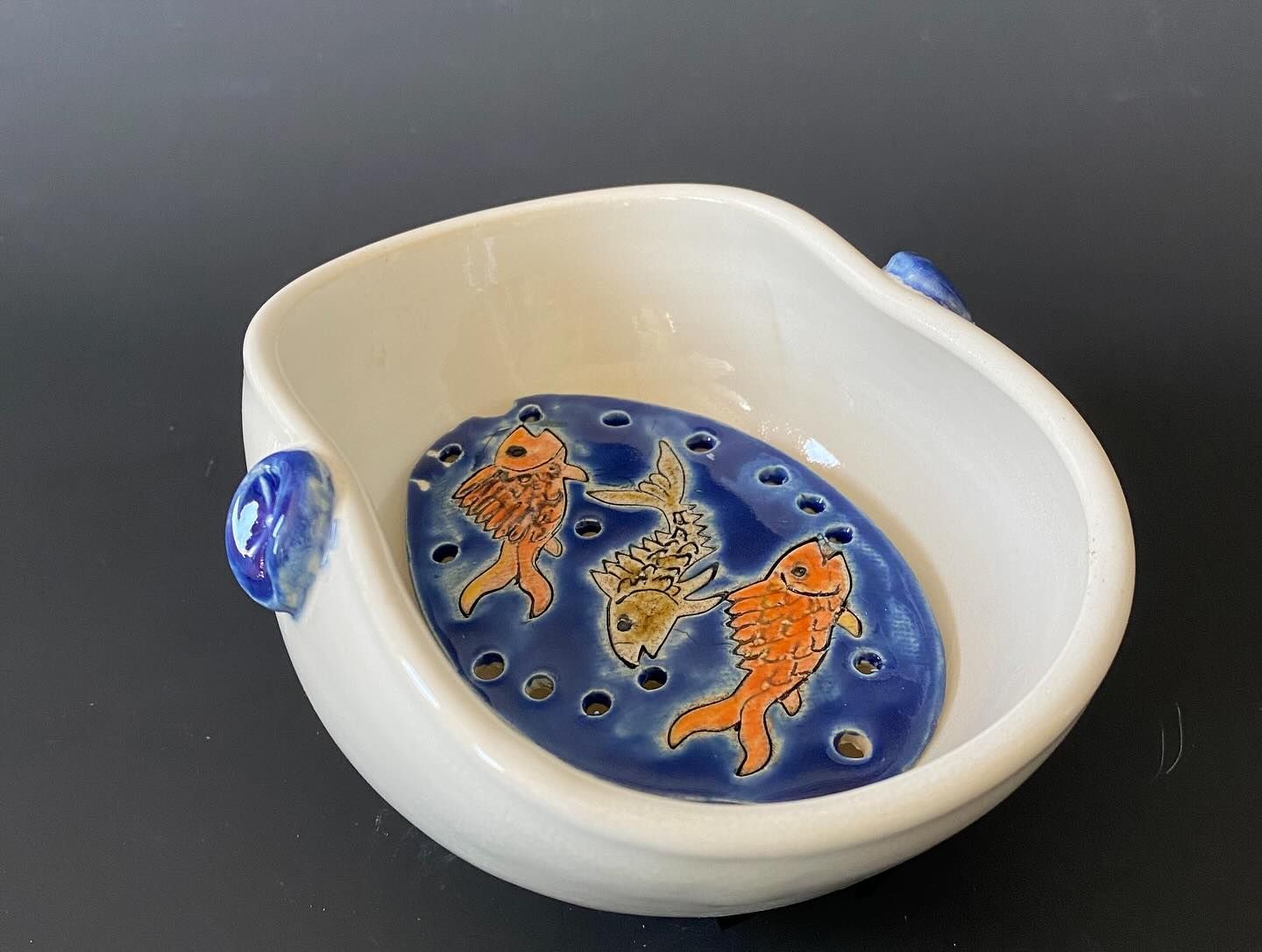 White ceramic bowl with a blue fish design in the center, featuring orange and yellow fish. Blue handles are on the sides.