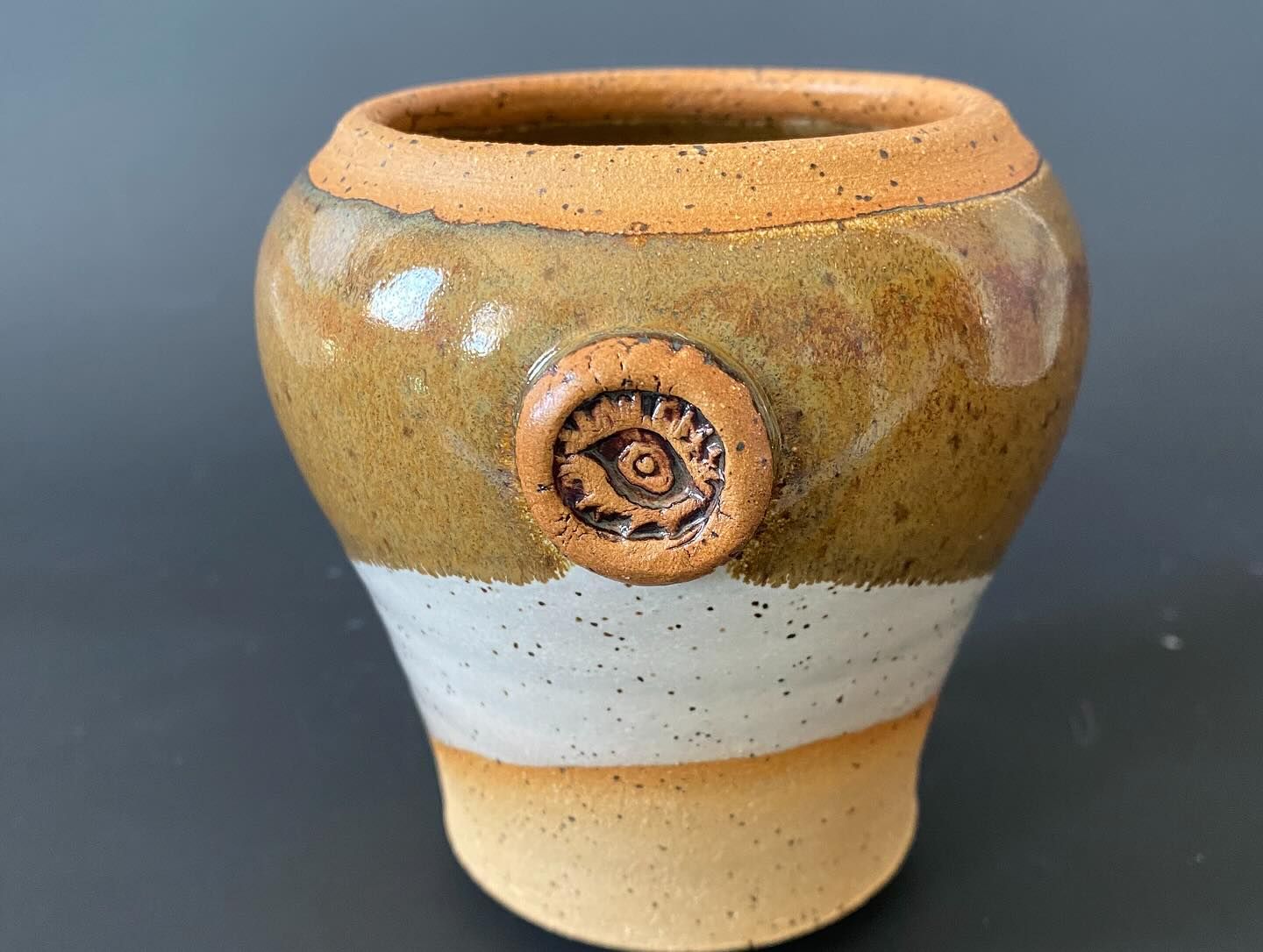 Ceramic vase with a brown top, white middle band, and yellow base; a brown eye-shaped decoration adorns the white band.