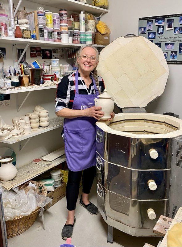 Woman in purple apron smiling, holding pottery near a kiln in a workshop.