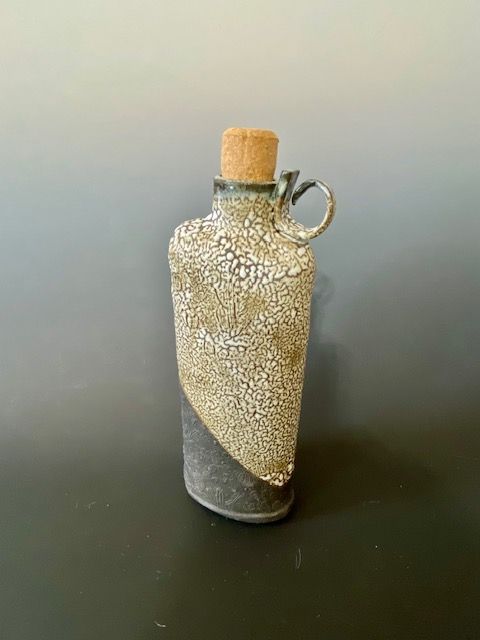 Ceramic bottle with a cork stopper and handle. The bottle is mostly tan with a dark gray base and a cracked glaze.