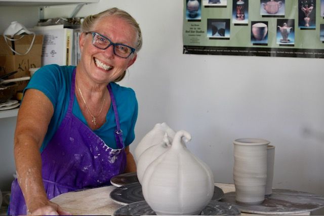 Woman smiling, wearing glasses and apron, working on ceramic art in a studio.