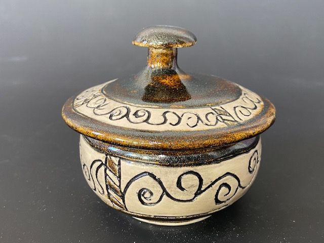 Ceramic lidded jar with dark brown top and rim, light-colored body with black line designs on a black surface.