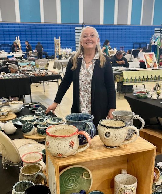 Woman at craft fair, standing by pottery display. Smiling, wearing black cardigan, grey hair. Indoor setting.