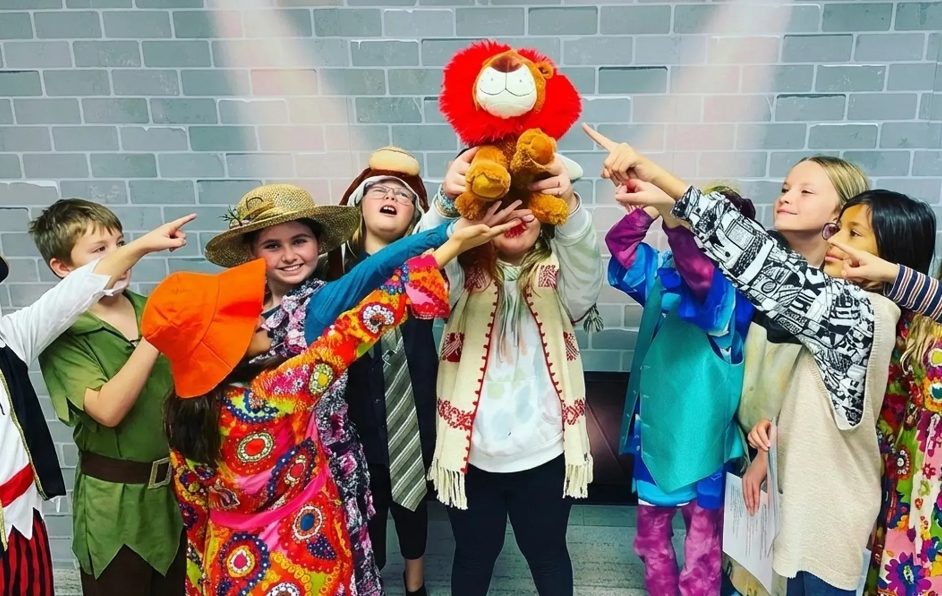 Children in costumes point at a plush lion, lit by spotlights against a brick wall.