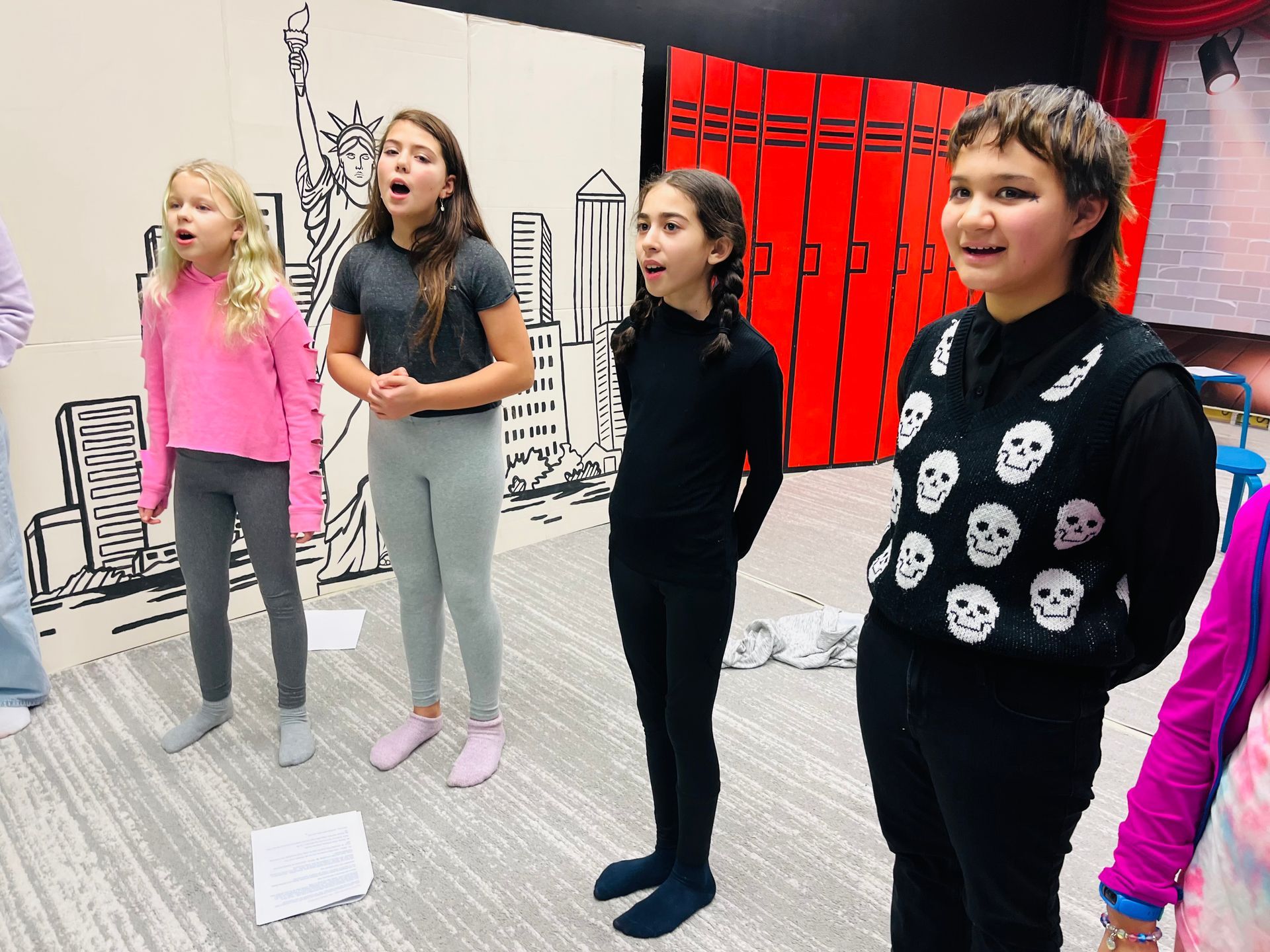 Children singing onstage, practicing, with city backdrop and red lockers.