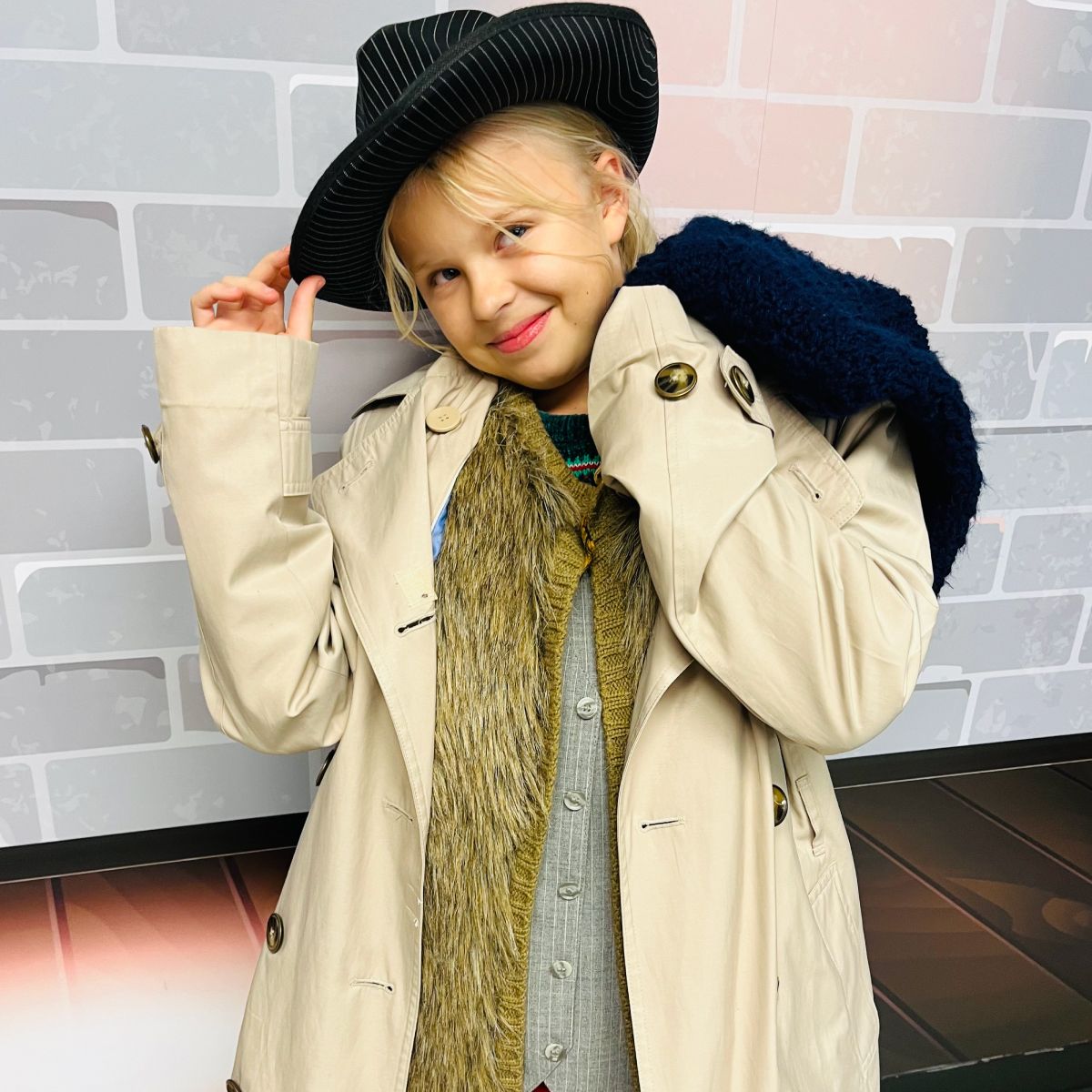 Girl in trench coat, fedora, and scarf smiles, posing against a brick wall background.