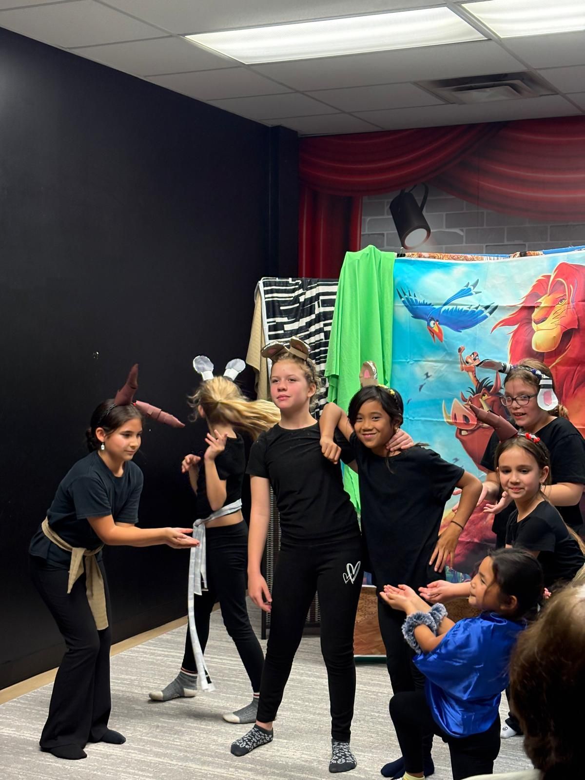 Children performing on stage, wearing costumes and acting. Black backdrop, colorful mural.