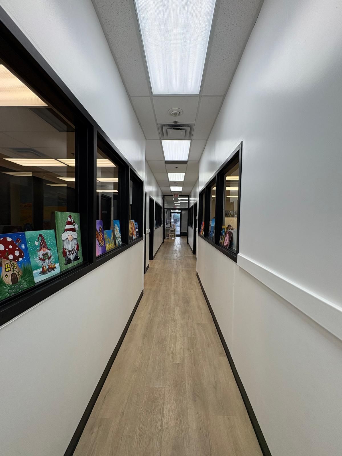 Long, narrow hallway with windows on both sides, artwork on display, and bright ceiling lights.