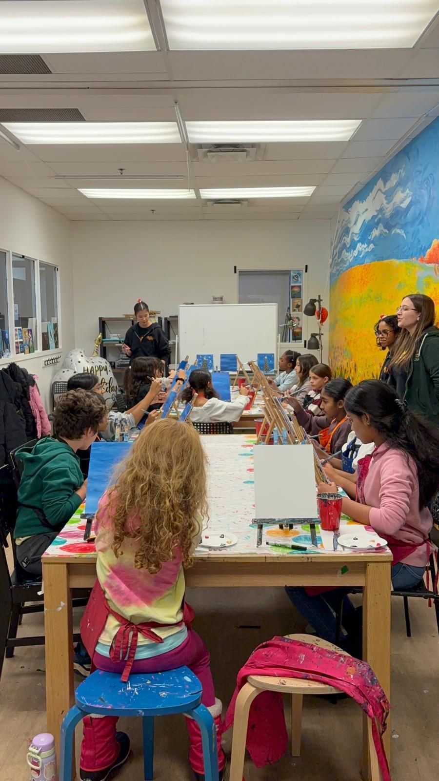 Children painting at tables in a classroom; an instructor leads, blue and white canvases are set up.