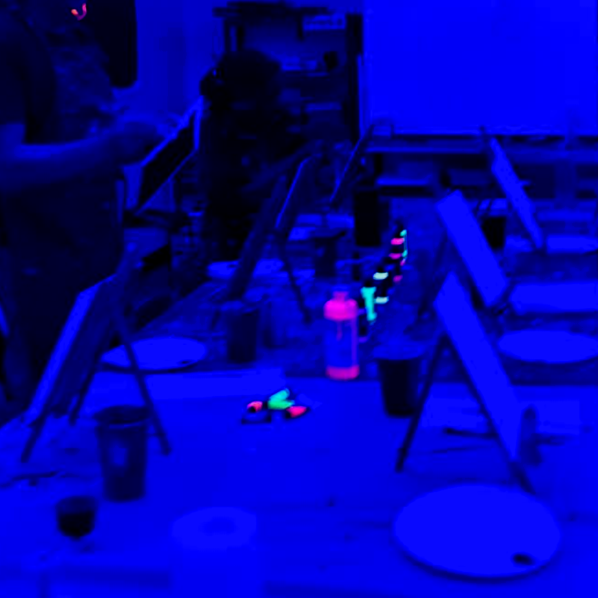 People painting at a table in a dimly lit room with glowing paint and easels.