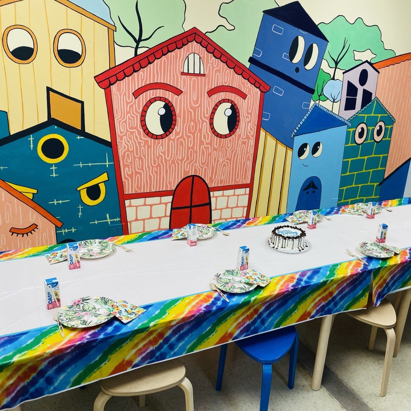 A table set for a party, in front of a cartoon cityscape mural. The table is decorated with rainbow colors.