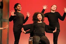 Three people in black clothing performing on a stage with red backdrop, arms outstretched.