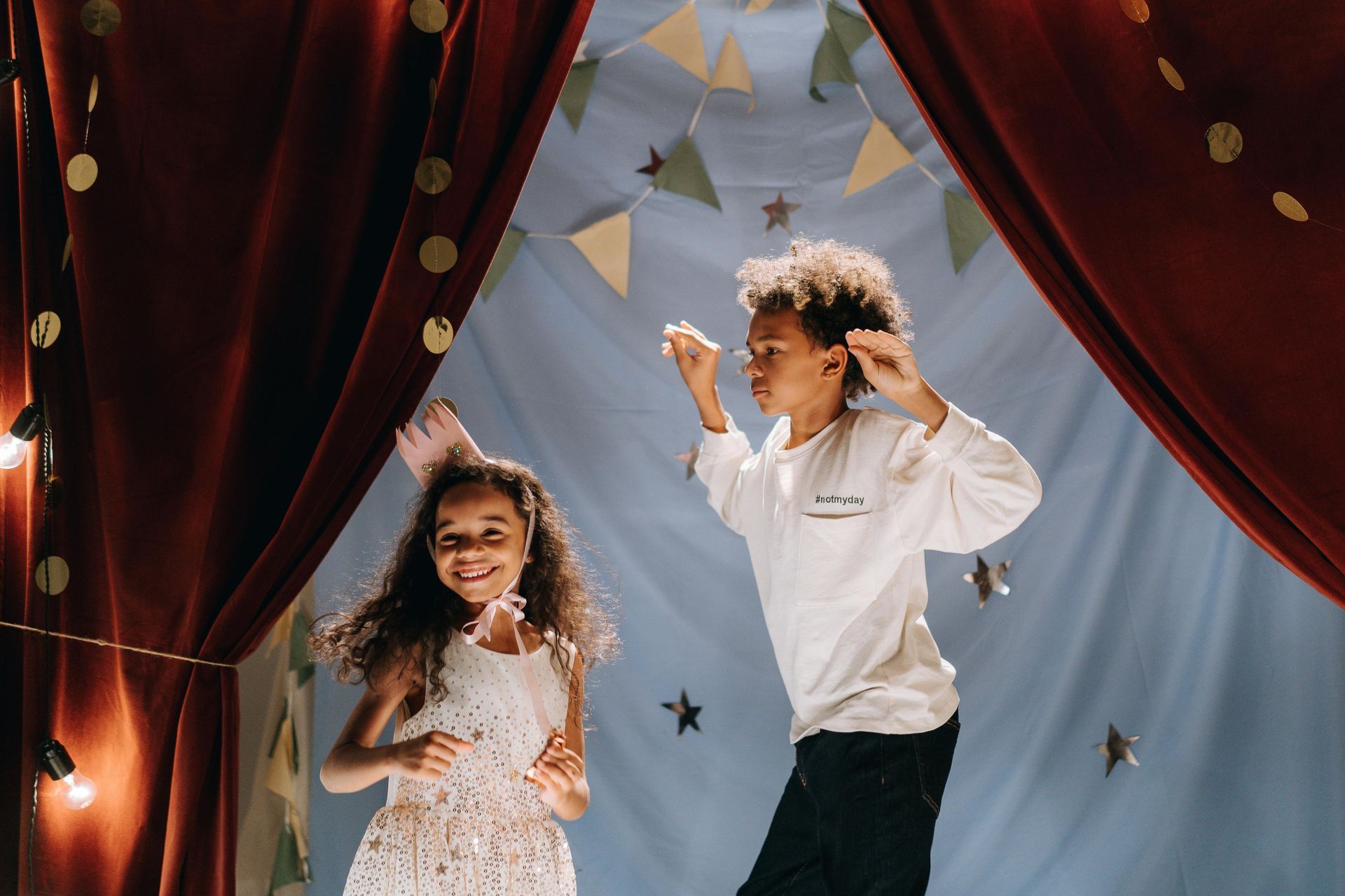 Two children dancing on a stage with red curtains and festive decorations; a girl in white and a boy in white.