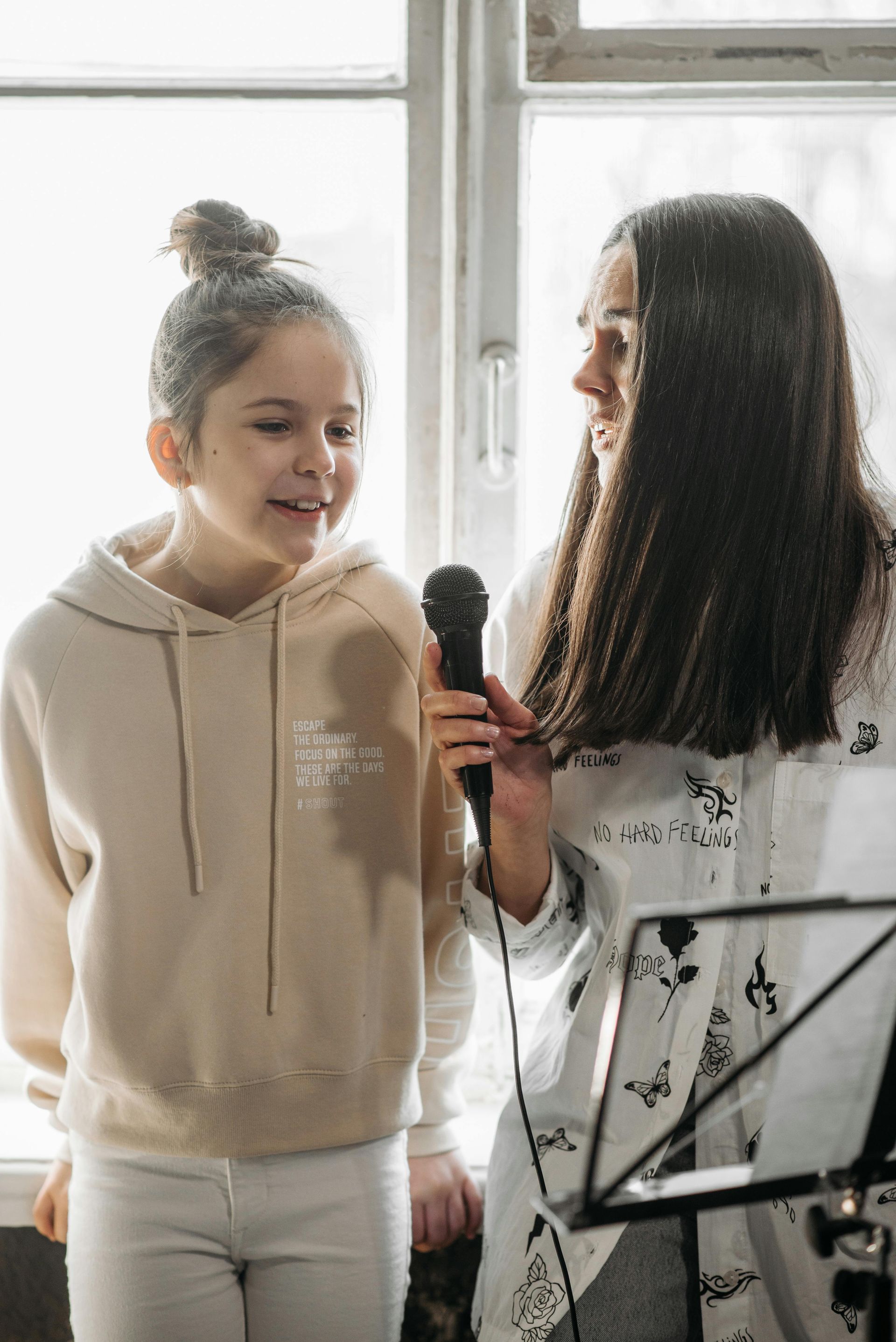 Woman holding microphone; girl smiles. Both are near a window. Music stand is on right.
