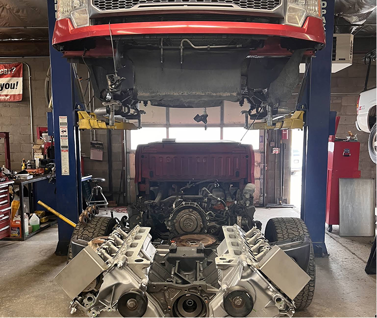 Car lift in auto shop with a red vehicle raised above a disassembled engine and transmission on the floor.