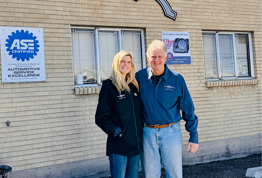 Two people standing in front of a brick building with an ASE sign, smiling outdoors.