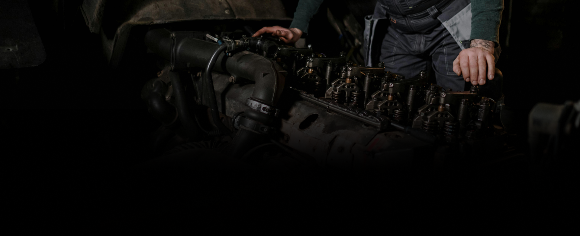 Dark scene with a person beside a black motorcycle, adjusting its handlebars or engine area.