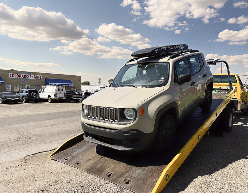 Silver Jeep on a tow truck ramp in a parking lot under a partly cloudy sky