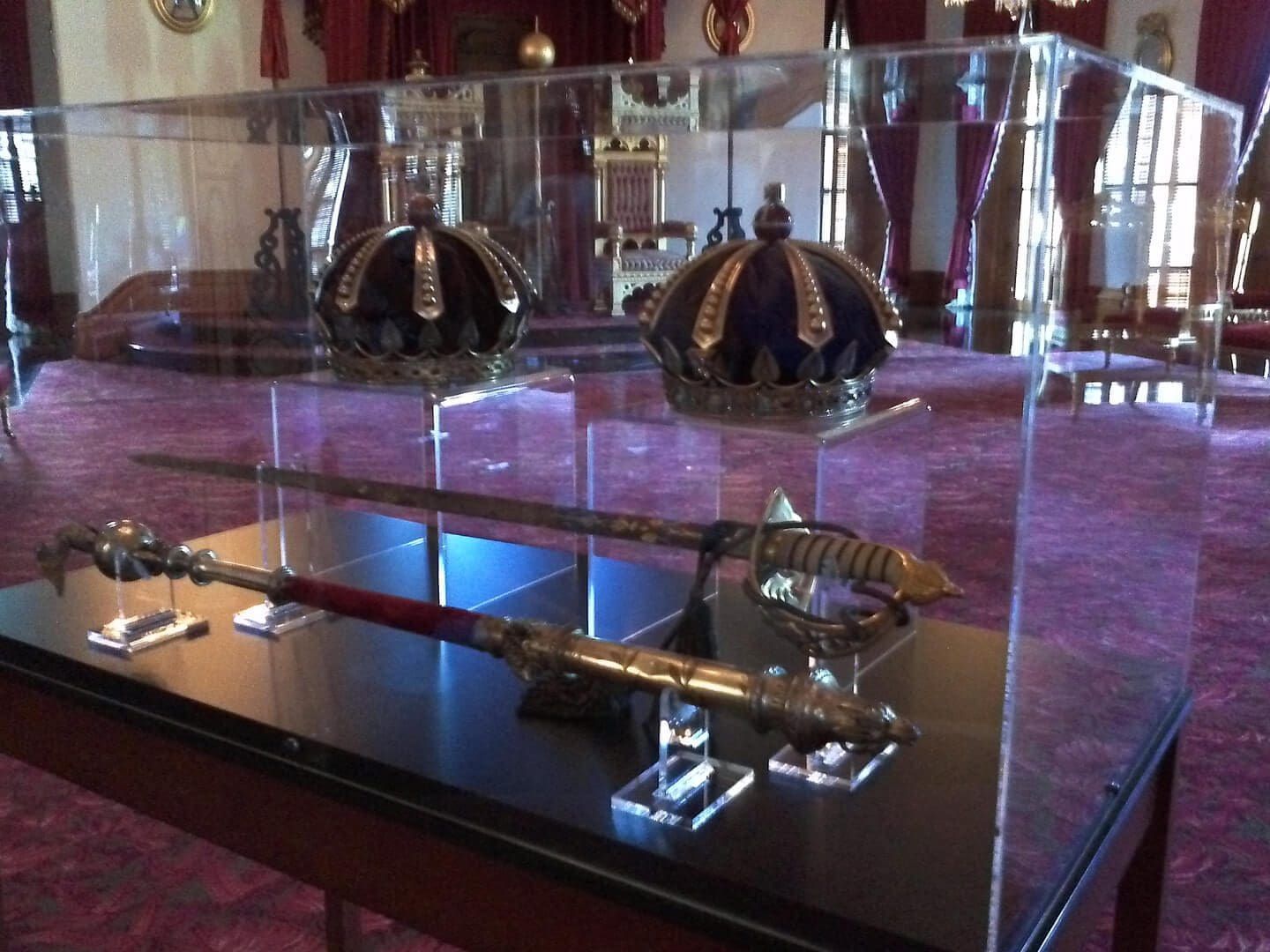 Display case with crowns, a sword, and a ceremonial mace on a reflective black surface in a room with red carpet.