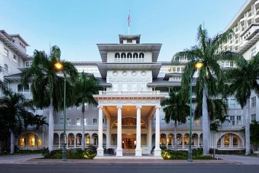White beachfront hotel with columns, palm trees, and flag.