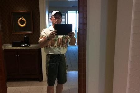 Man in Hawaiian shirt, shorts, and hat taking a selfie in a mirror.
