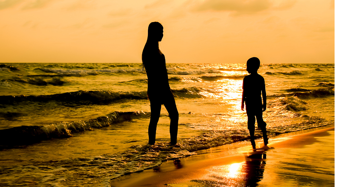 A woman and a child are standing on a beach at sunset.