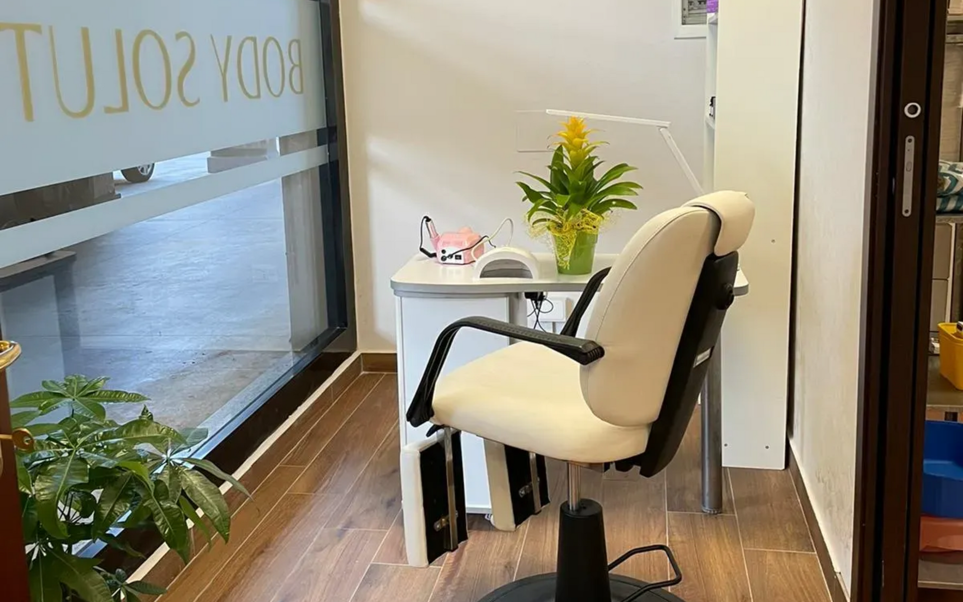 Postazione per manicure da salone con sedia e scrivania bianche, piante e porta in vetro con logo.