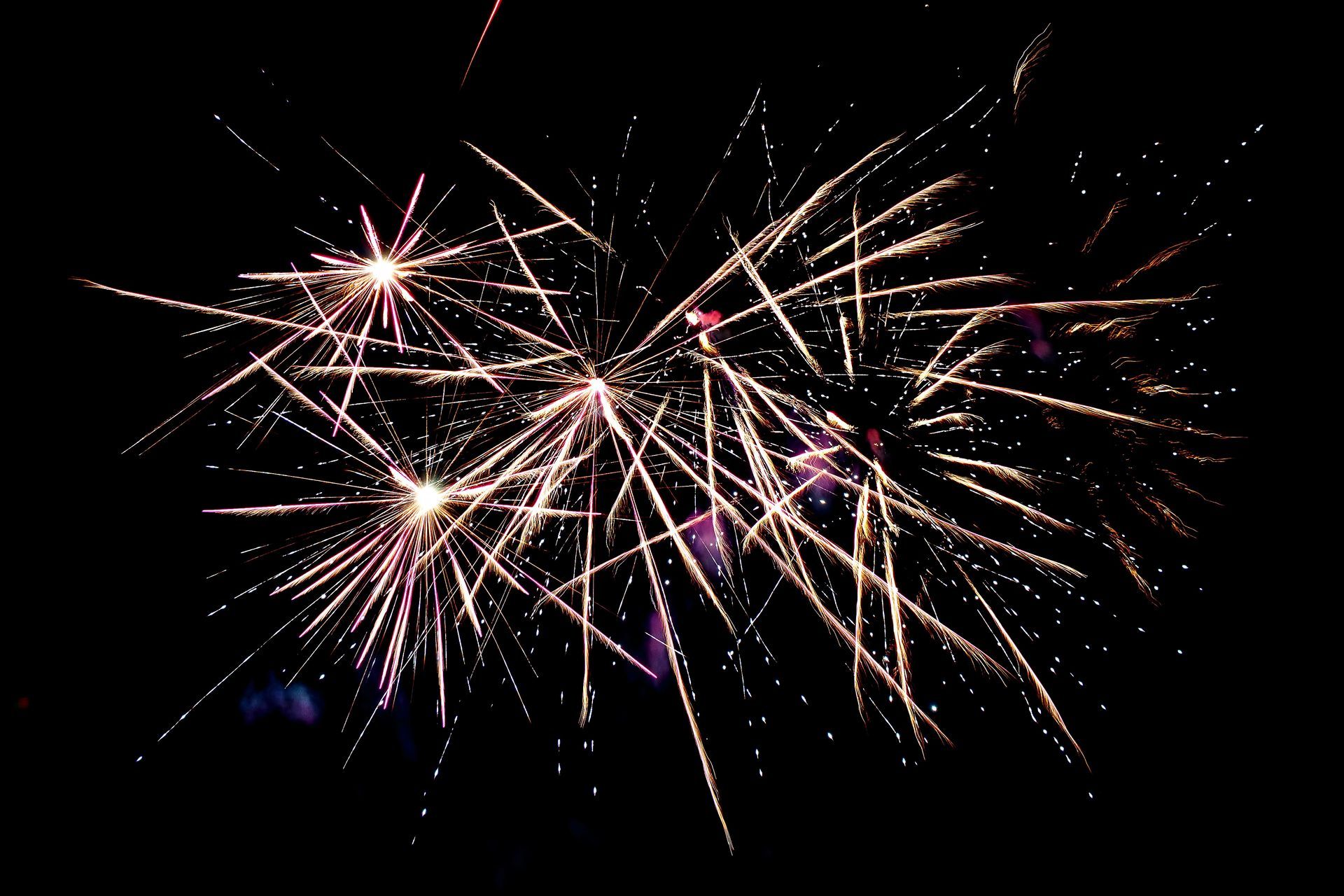 Fuochi d'artificio che esplodono nel cielo notturno, con scie luminose bianche e rosa su uno sfondo nero.