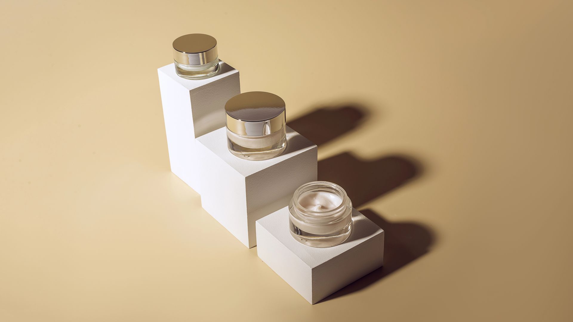 Tre barattoli di crema per la cura della pelle con coperchi dorati sono appoggiati su blocchi geometrici bianchi