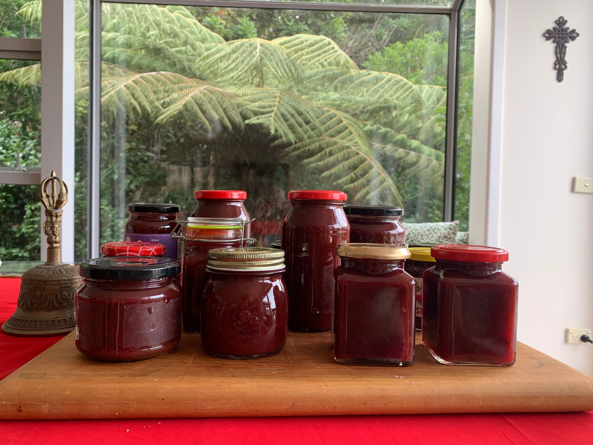 Jars of reddish-brown jam with various lids, on a wooden board in front of a window with green foliage.