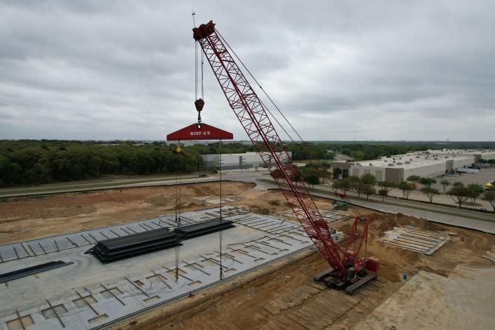 Red crane lifting a large red concrete slab at a construction site under a cloudy sky