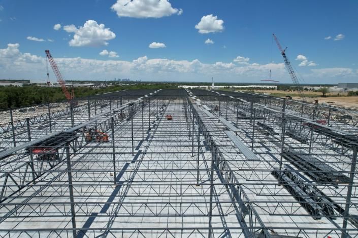 Steel building frame under construction on a large site, with cranes and a bright blue sky with clouds.