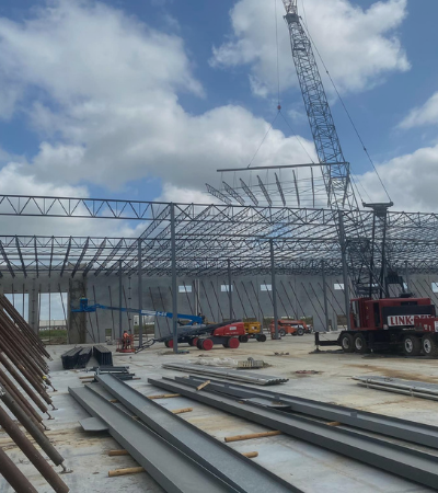 Steel-frame building under construction with a tall blue crane and stacked beams in the foreground.