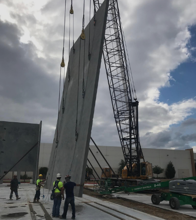 Workers and a crane lifting large concrete panels at an industrial site under a cloudy sky