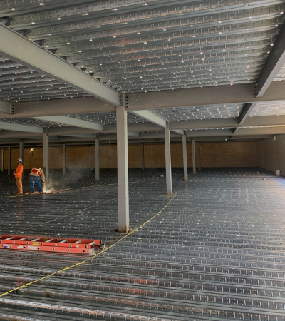 Steel-framed building interior under construction, with workers on the left and exposed floor decking throughout.