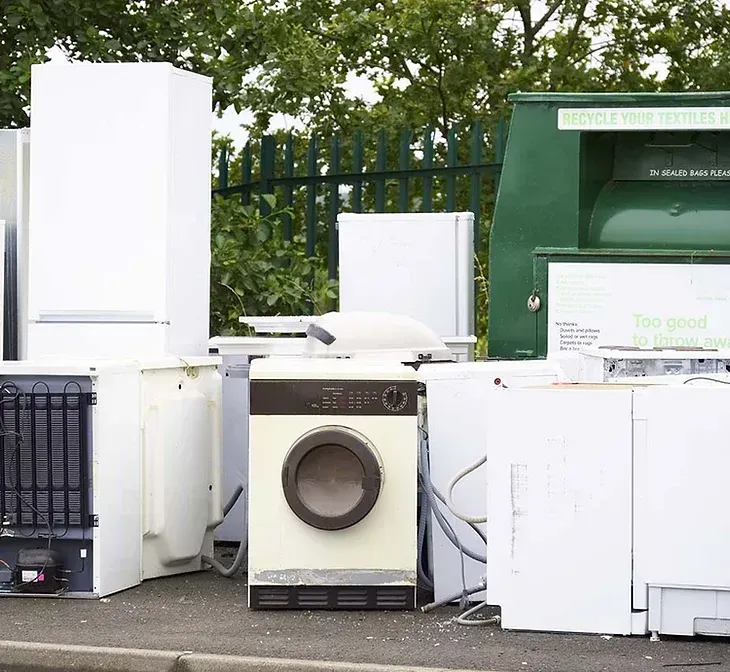 Discarded appliances and household items piled on a street beside a green dumpster.