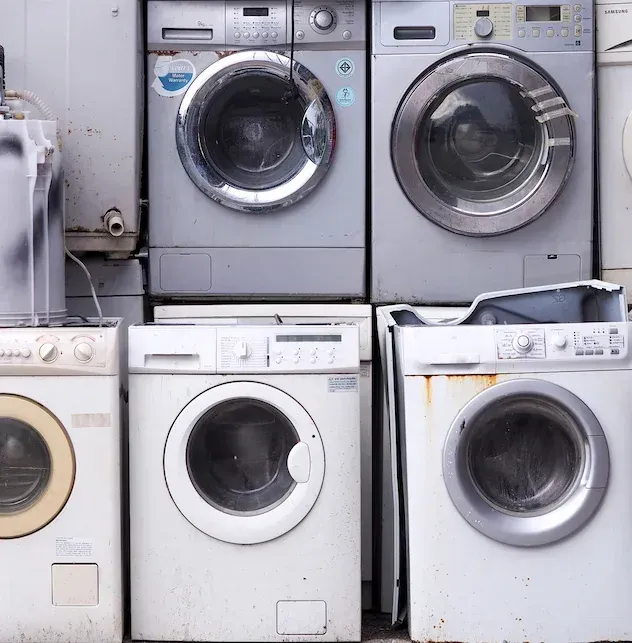 Stacked front-loading washing machines in a laundromat, mostly white and gray, some worn and stained