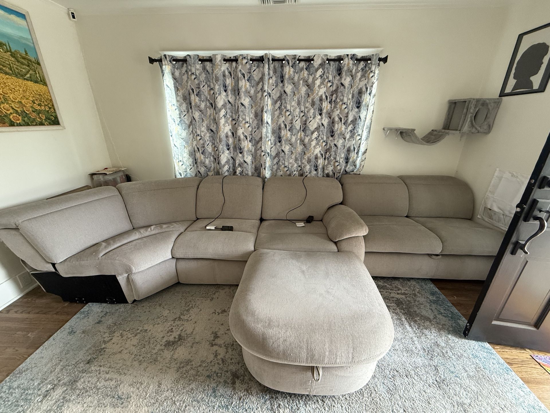 Beige sectional sofa with ottoman in a living room, with patterned curtains and wall shelves.